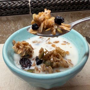 May include: A light blue ceramic bowl filled with granola and milk. A spoonful of granola with blueberries is held above the bowl. The granola appears to have oats, seeds, and dried fruit.