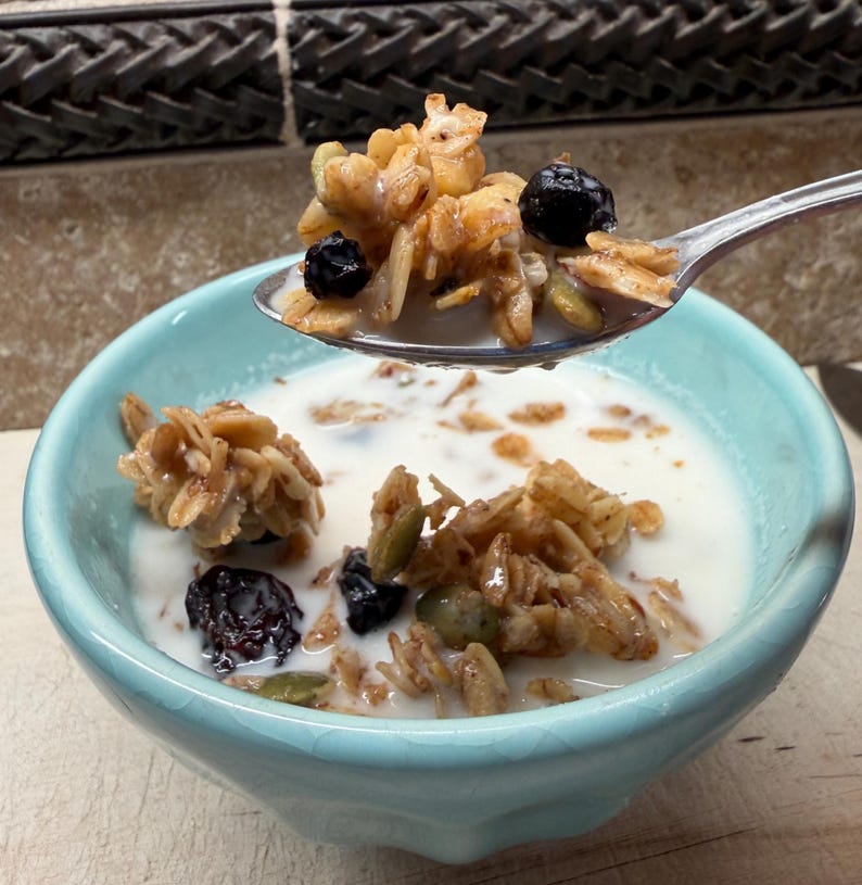 May include: A light blue ceramic bowl filled with granola and milk. A spoonful of granola with blueberries is held above the bowl. The granola appears to have oats, seeds, and dried fruit.