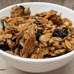 May include: A white ceramic bowl filled with homemade granola. The granola is a mix of golden-brown oats, nuts, seeds, and dried fruit, including dark raisins and cranberries. The bowl is set against a neutral background.