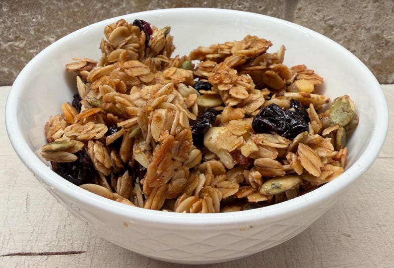 May include: A white ceramic bowl filled with homemade granola. The granola is a mix of golden-brown oats, nuts, seeds, and dried fruit, including dark raisins and cranberries. The bowl is set against a neutral background.