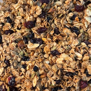 May include: Close-up of a mix of granola with oats, nuts, seeds, and dried berries. The granola has a golden-brown color, with visible pieces of almonds, pumpkin seeds, and dried cranberries and blueberries. A healthy breakfast or snack.