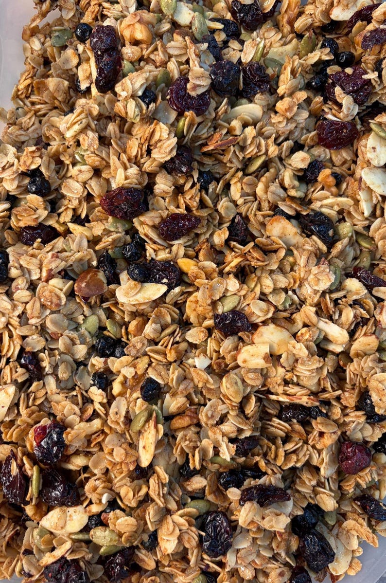 May include: Close-up of a mix of granola with oats, nuts, seeds, and dried berries. The granola has a golden-brown color, with visible pieces of almonds, pumpkin seeds, and dried cranberries and blueberries. A healthy breakfast or snack.
