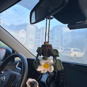 May include: A crocheted flower car air freshener hanging from the rearview mirror of a car. The flower is white with a yellow center and green leaves. The air freshener is hanging from a brown string.