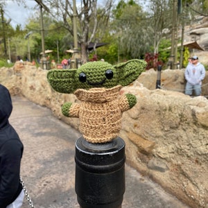 May include: A green and brown crocheted plush toy of a character from the Star Wars franchise, standing on a black and brown metal post. The toy has large eyes and a smiling expression.