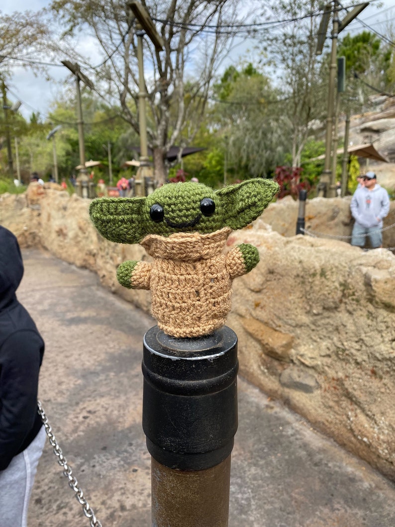 May include: A green and brown crocheted plush toy of a character from the Star Wars franchise, standing on a black and brown metal post. The toy has large eyes and a smiling expression.