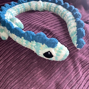 May include: A plush, crocheted sea serpent toy in shades of blue, white, and teal. The toy features a dark blue spine and large, black eyes. The serpent is lying on a purple corduroy surface.