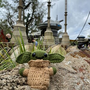 May include: A green and brown crocheted plush toy of a popular character from the Star Wars franchise, sitting on a rock in front of a Star Wars themed background.