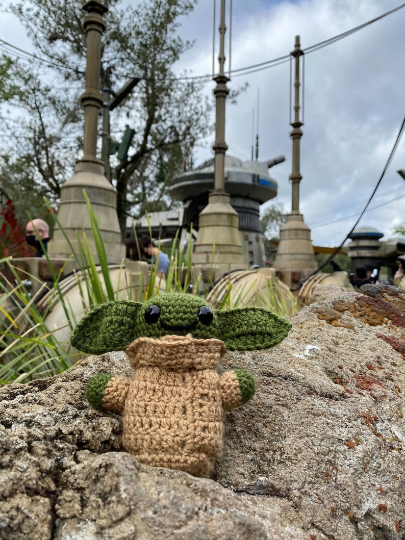 May include: A green and brown crocheted plush toy of a popular character from the Star Wars franchise, sitting on a rock in front of a Star Wars themed background.