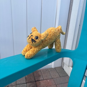 May include: A handmade, golden-yellow crochet cat toy with black facial details, posed on a teal blue surface. The plush cat has a playful, outstretched posture, with its paws extended. The toy is made of a textured yarn.