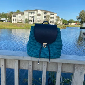 May include: Teal blue woven backpack with a black leather flap and gold buckle. The backpack is sitting on a wooden railing with a lake and buildings in the background.