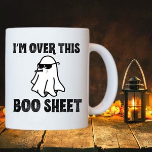 May include: White ceramic coffee mug with a black and white illustration of a ghost wearing sunglasses and a cigarette. The text "I'M OVER THIS BOO SHEET" is printed on the mug.