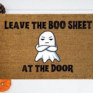 May include: A brown doormat with the text "Leave the Boo Sheet at the Door" in black. A cartoon ghost with crossed arms is in the center of the mat.