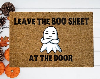 May include: A brown doormat with the text "Leave the Boo Sheet at the Door" in black. A cartoon ghost with crossed arms is in the centre of the mat.