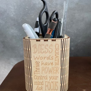 May include: A wooden desk tidy containing pens, scissors, and a ruler. The organiser has a laser-cut design with the words "Bless, Words have POWER, Control your mind, Ask FOCUS". The organiser is made of light-coloured wood.