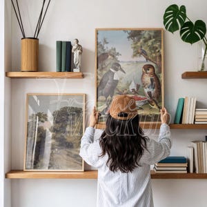 May include: A person holding a framed vintage bird print with a brown cap. The print features a detailed illustration of birds in a natural setting. The room has wooden shelves with books, a statue, and decorative items.