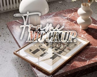 May include: A rectangular wooden picture frame containing a geometric print with a grid pattern, resting on a red marble table. A white mushroom-shaped lamp and a white ceramic vase are nearby. The text "Frame MOCKUP." and "PSD" are visible.