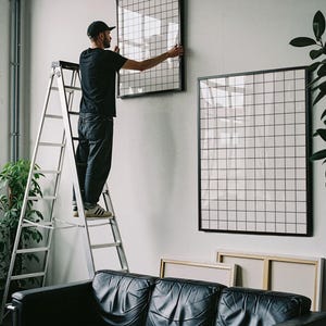 Pode incluir: Duas grandes pe&ccedil;as de arte de parede retangulares com um padr&atilde;o de grade preta sobre fundo branco, emolduradas com bordas pretas finas. Uma pe&ccedil;a est&aacute; montada na parede, enquanto a outra est&aacute; posicionada abaixo, encostada na parede perto de um sof&aacute; preto.