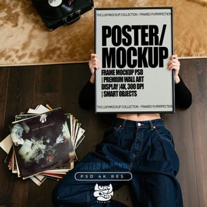 May include: A black framed poster mockup held by a person wearing blue jeans and a black long-sleeve shirt. The poster has the text "POSTER/MOCKUP" and other details. A stack of vinyl records and a record player are also visible.