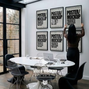 Puede incluir: Un moderno entorno de oficina con una mesa ovalada blanca, sillas de alambre y un portátil. Se exhiben seis carteles enmarcados con el texto "POSTER/MOCKUP" en la pared. Una persona está ajustando uno de los carteles. La habitación tiene ventanas grandes.