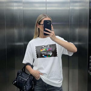 Puede incluir: Una camiseta blanca de manga corta con un gr&aacute;fico de patr&oacute;n de prueba de televisi&oacute;n dentro de un rect&aacute;ngulo de cuadr&iacute;cula negra. La prenda se combina con vaqueros de mezclilla gris oscuro y un bolso de cuero negro con herrajes plateados.