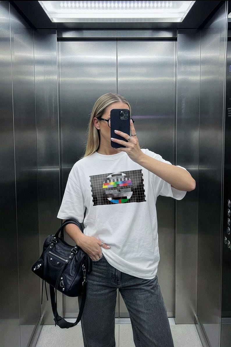 Puede incluir: Una camiseta blanca de manga corta con un gr&aacute;fico de patr&oacute;n de prueba de televisi&oacute;n dentro de un rect&aacute;ngulo de cuadr&iacute;cula negra. La prenda se combina con vaqueros de mezclilla gris oscuro y un bolso de cuero negro con herrajes plateados.
