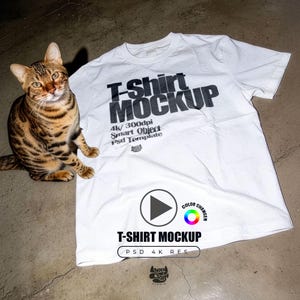 Puede incluir: Una maqueta de camiseta blanca con el texto "T-Shirt MOCKUP" en negro. Un gato bengalí con marcas marrones y negras está sentado a la izquierda de la camiseta. La camiseta también tiene el texto "4K/300dpi Smart Object Psd Template".