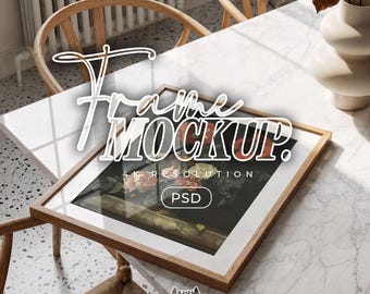 May include: A rectangular wooden picture frame mockup resting on a white marble table. The frame contains a dark floral art print. Overlaid text reads Frame MOCKUP. 4K RESOLUTION PSD. A beige ceramic vase sits nearby on the table surface.