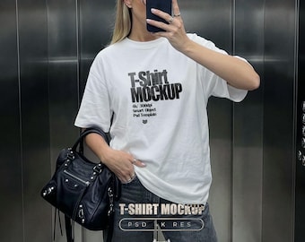 May include: A white short-sleeved t-shirt featuring the black printed text T-Shirt MOCKUP, 4k/ 300dpi, Smart Object, Psd Template. The shirt is paired with dark grey jeans and a black leather handbag with silver hardware and multiple zippered pockets.