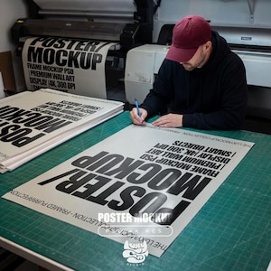 May include: A person wearing a maroon cap and black hoodie is working on a large poster mockup. The poster features bold black text that reads "POSTER MOCKUP" and includes details like "FRAME MOCKUP PSD" and "PREMIUM WALL ART". The scene takes place in a print studio.