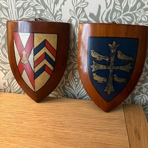 May include: Two wooden wall plaques shaped like heraldic shields. The left shield features a red saltire with a gold scallop shell and a gold field with blue and red chevrons. The right shield displays a blue field with a gold cross and four birds.