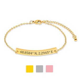 May include: Gold-colored bracelet with a curved bar engraved with coordinates: 48.8584° N, 2.2945° E. The bracelet has a delicate chain and a lobster clasp closure. Three color swatches are shown below the bracelet: yellow, gray, and pink.