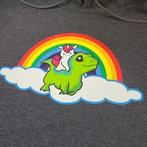 May include: A gray hoodie with a green dinosaur riding a unicorn on a rainbow over a white cloud. The unicorn has a pink horn and a white mane.