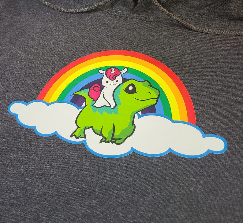 May include: A gray hoodie with a green dinosaur riding a unicorn on a rainbow over a white cloud. The unicorn has a pink horn and a white mane.