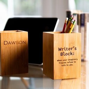Może przedstawiać: Dwa drewniane stojaki na ołówki, jeden z wygrawerowanym imieniem "DAWSON" z przodu, a drugi z tekstem "Writer's Block: When your imaginary friends refuse to talk to you."