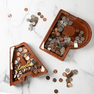 May include: Two wooden money boxes shaped like the letters A and P, with a clear plastic front to see the coins inside. The A money box has the name Amelia written on it in gold lettering. Both money boxes are filled with coins.