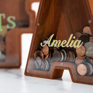 May include: Wooden piggy bank shaped like the letter A with a clear plastic front. The piggy bank is filled with coins and has the name "Amelia" written on the front in gold lettering.