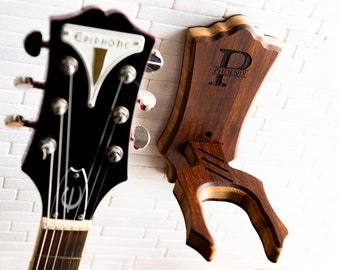 May include: A dark wood guitar stand with a personalized engraving that reads "P Phoenix". The stand is next to the headstock of a black Epiphone guitar. The guitar stand is designed to hold a guitar.