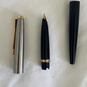May include: A disassembled fountain pen with a silver cap and gold clip, a black barrel with a gold band, and a black cap. The nib is gold-colored. The pen is lying on a white surface.