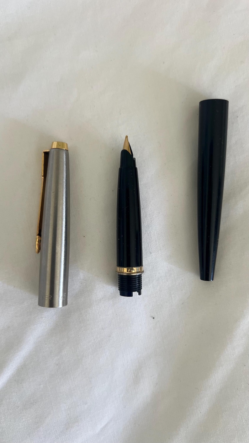 May include: A disassembled fountain pen with a silver cap and gold clip, a black barrel with a gold band, and a black cap. The nib is gold-colored. The pen is lying on a white surface.