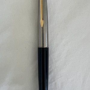 May include: A vintage fountain pen with a silver barrel and a black lower body. The pen features a gold-colored arrow-shaped clip and nib. The pen is a classic writing instrument.