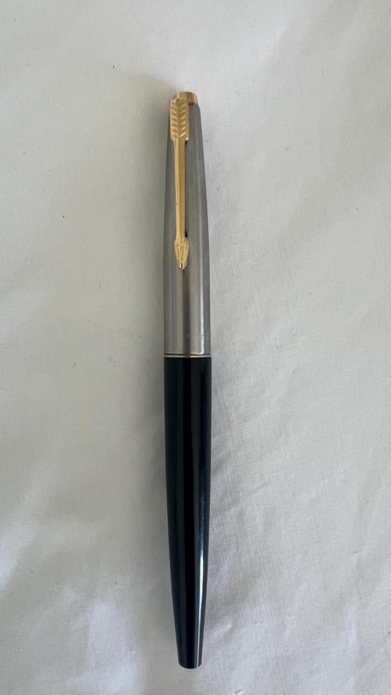 May include: A vintage fountain pen with a silver barrel and a black lower body. The pen features a gold-colored arrow-shaped clip and nib. The pen is a classic writing instrument.