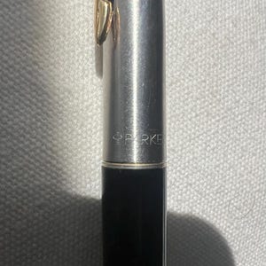 May include: A close-up of a Parker pen. The pen has a silver body with a gold-colored clip. The lower portion of the pen is black. The word "PARKER" is printed on the pen's body.