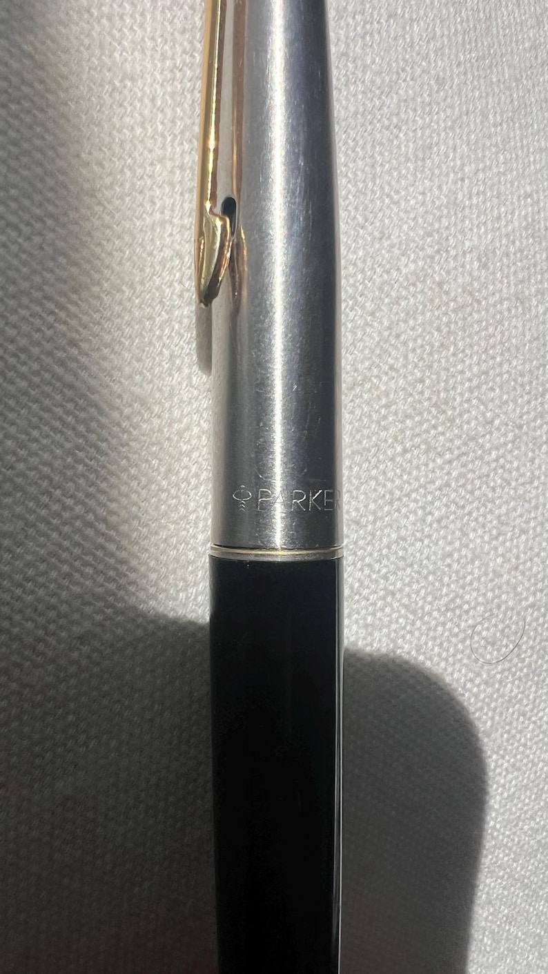 May include: A close-up of a Parker pen. The pen has a silver body with a gold-colored clip. The lower portion of the pen is black. The word "PARKER" is printed on the pen's body.