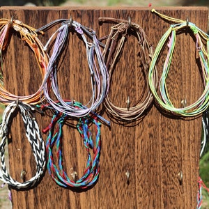 May include: A wooden board with colorful string bracelets hanging from hooks. The bracelets are made of various colors, including blue, green, red, white, and black.