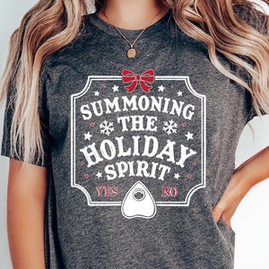 May include: Dark gray t-shirt with a white and red graphic. The graphic reads "Summoning the Holiday Spirit" with a bow, snowflakes, and a planchette. The shirt also has the words "Yes" and "No".