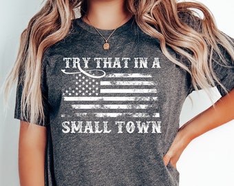 Puede incluir: Una camiseta gris oscuro con una bandera estadounidense blanca desgastada y el texto "Try that in a small town".