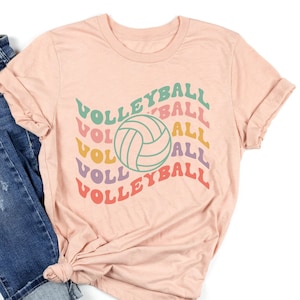 Könnte beinhalten: Ein pfirsichfarbenes T-Shirt mit dem Wort "VOLLEYBALL" in einem wellenförmigen Design, mit einer Volleyball-Grafik. Das Shirt ist mit blauen Jeans kombiniert. Das T-Shirt ist unten geknotet.
