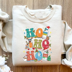May include: Cream-colored sweatshirt with a colorful "HO HO HO" graphic. The design incorporates Christmas elements like Santa hats, a snowman, and a gingerbread man, along with medical-themed imagery. The sweatshirt has a crew neck and long sleeves.