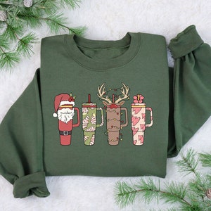 May include: An olive green sweatshirt with four Christmas-themed coffee cup designs. The cups feature Santa, candy canes, reindeer, and gifts. The cups are in red, green, brown, and pink, with a white background. The sweatshirt has long sleeves.