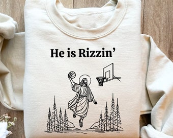 May include: A beige sweatshirt with a black line drawing of a person playing basketball with the text "He is Rizzin'" printed on the front.